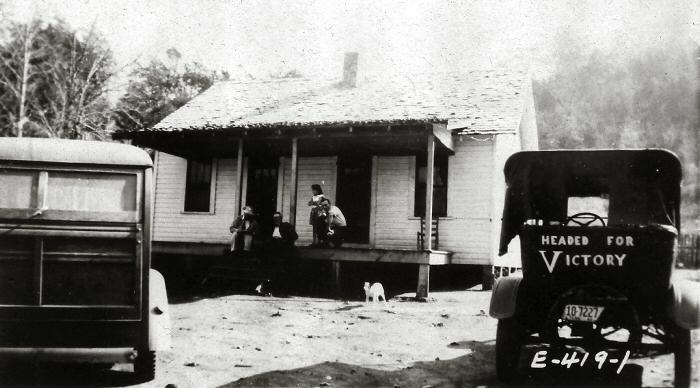 Old dwelling with a front porch with people and dog and two cars in front of the house